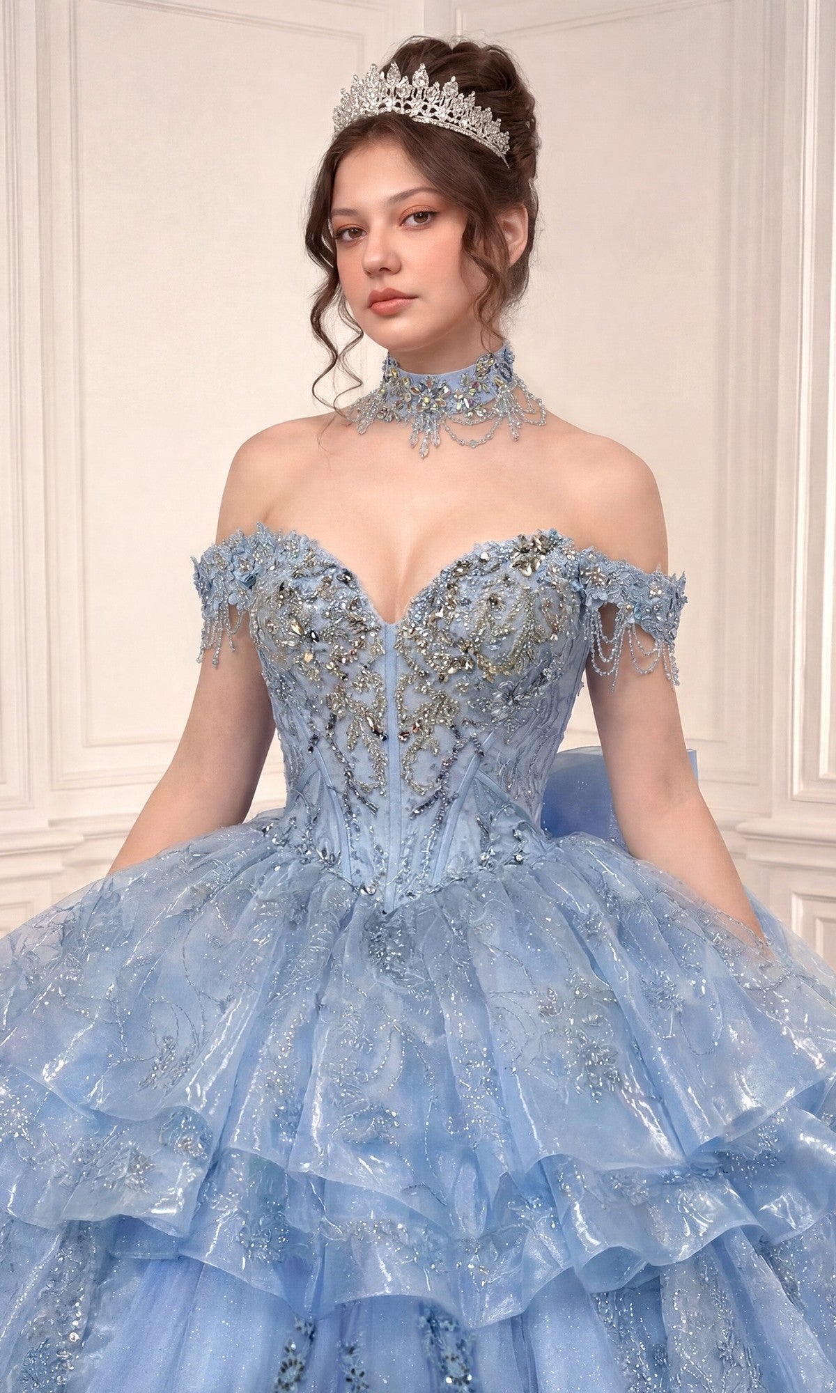 A woman wearing Ladivine’s Quinceanera Dress 15742, a light blue off-the-shoulder beaded gown paired with a lace choker and tiara, stands against a light-paneled background.